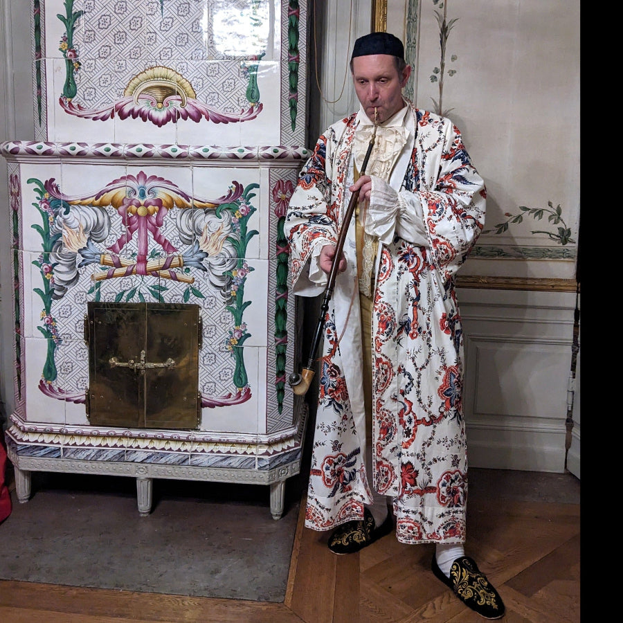 Man in traditional attire standing in a room with decorative tiles and a fireplace.