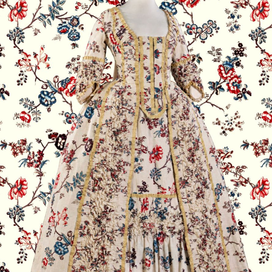 A long Victorian-style gown displayed on a mannequin against a matching floral wallpaper background. The dress has a warm beige base with an all-over multicolor floral print in red, blue, and green, accented by gold trim along the neckline, front opening, and sleeve edges.